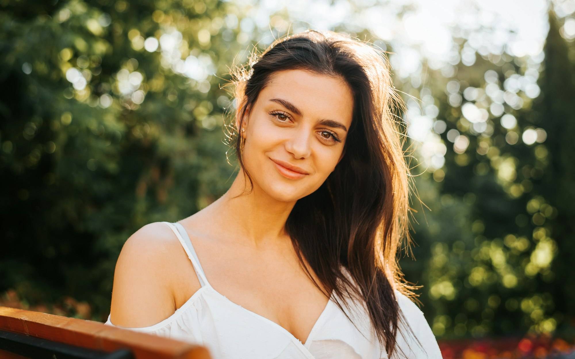 Portrait pretty young brunette woman in white light dress with gentle smile relaxing outdoors and